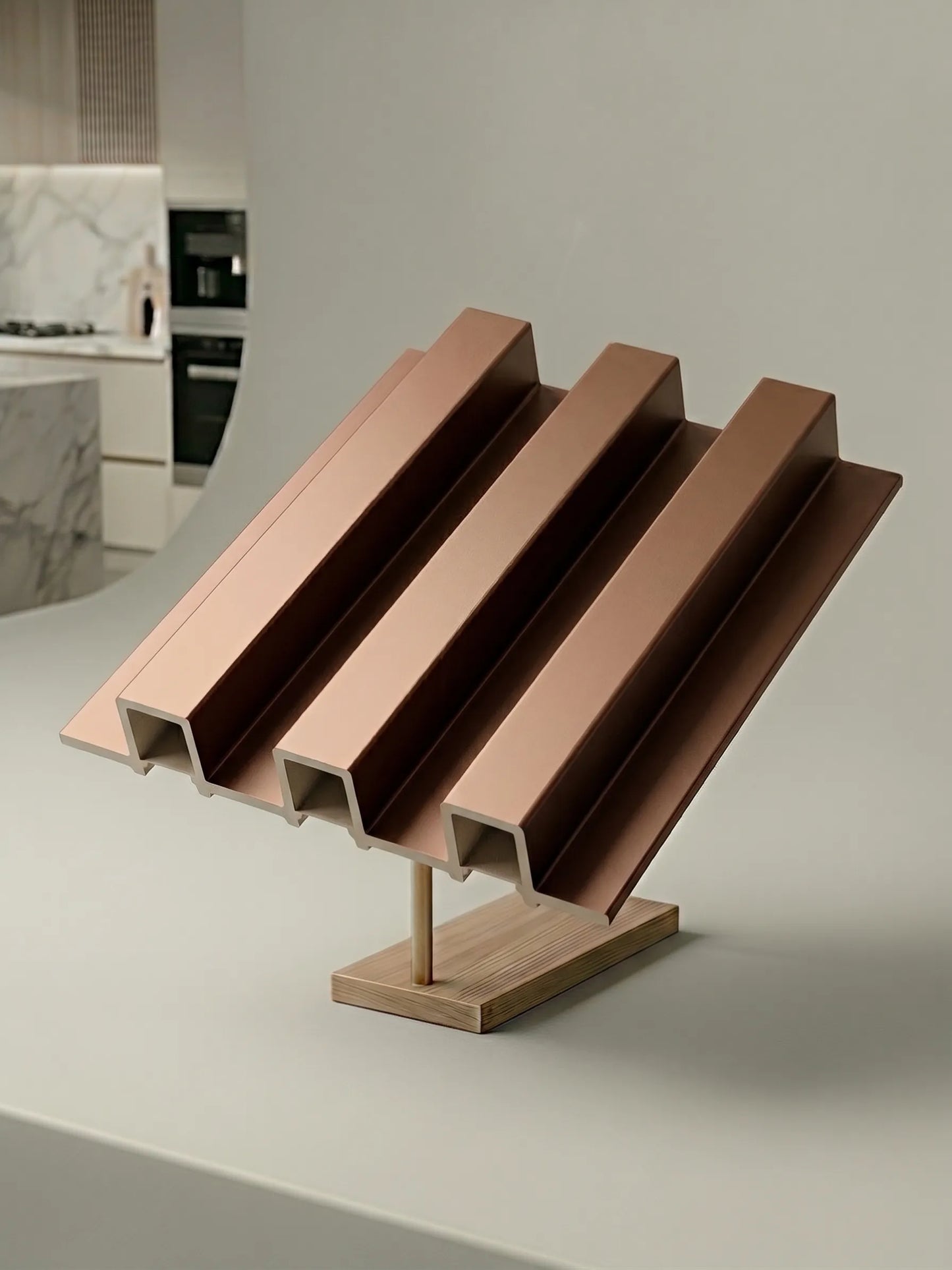 A modern decorative object with four parallel angled metallic rose-gold ridges mounted on a light wood base, photographed on a clean white surface with a softly blurred kitchen interior in the background.