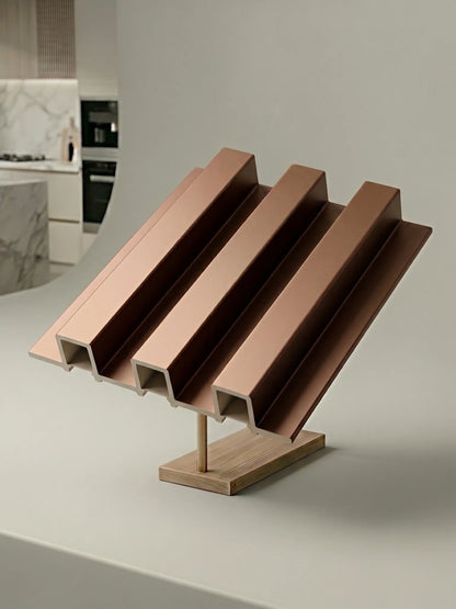 A modern decorative object with four parallel angled metallic rose-gold ridges mounted on a light wood base, photographed on a clean white surface with a softly blurred kitchen interior in the background.